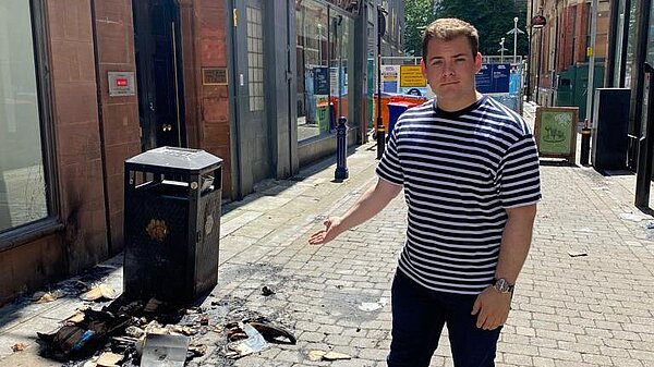 Image of Luke, pointing at a pile of burned cardboard beside a litter bin.