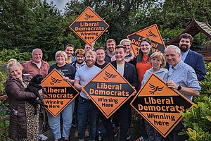 A diverse group of Liberal Democrat campaigners stand outside surrounded by trees and bushes, holding vibrant orange diamond signs featuring the recognisable Liberal Democrat logo and taglines.