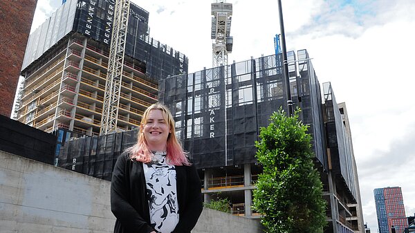 Chris Northwood with blonde and light pink hair stands in front of a construction site with several cranes and a high-rise building under construction, wrapped in netting branded with the name "RENAKER." She is smiling and wears a black cardigan over a patterned top.