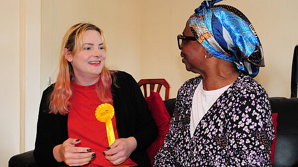Chris Northwood sits on a sofa wearing a Liberal Democrat rosette. She is in conversation with a local resident.