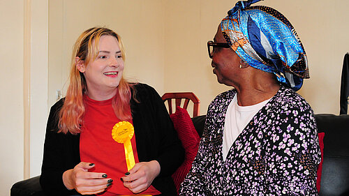 Chris Northwood sits on a sofa wearing a Liberal Democrat rosette. She is in conversation with a local resident.