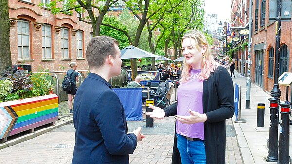 A woman with light blonde and pink hair is engaged in conversation with a man on a bustling street lined with brick buildings and outdoor seating. The woman is smiling, wearing a black cardigan over a purple top, and the man is dressed in a dark blazer.